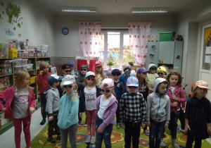 Dzieci ustawione w rozsypce na dywanie. Każde w bluzie sportowej, na głowach czapki z daszkiem u niektórych na oczach okulary słoneczne.